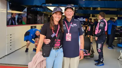 Lucy Wallduck And older man and young woman wearing VIP passes stand in front of a Formula 1 garage labelled. Team members are visible inside working on equipment and cars.