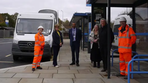 Six people are stood posing on a pavement in front of a bus stop. The two people on both ends, a man and a woman, are wearing orange hi vis jackets.