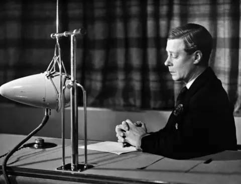 The then-Duke of Windsor sitting at a desk with a large microphone in front of him for a broadcast to the Empire