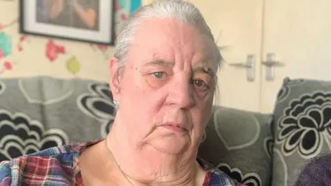 Phil Cunliffe/BBC Ann Bell, who has grey hair and a chequered blouse, sits on a couch She is looking into the camera with a sombre expression. 