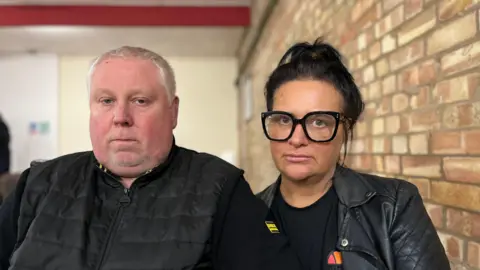 Abigail Jaiyeola/BBC Mark and Caroline Willgoose: a man and a woman both wearing black jackets and staring into the camera. Mark has short white hair, while Caroline has bobbed black hair and is wearing thick-rimmed spectacles. A brick wall is over Caroline's left shoulder.