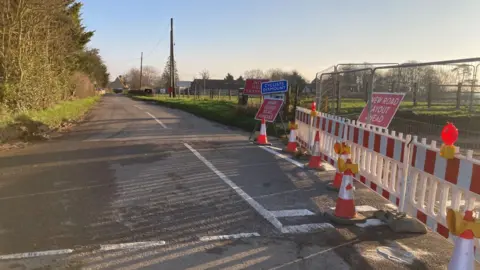 A road, in a village, that is closed. You can see a house in the distance, to the left, trees and a road that is open, next to a road that is closed, complete with metal and plastic barriers and cones.