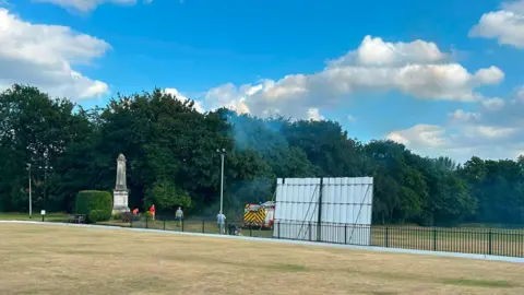 Stockport Cricket Club a fire engine, in the distance, is parked next to a bush. Smoke is coming from the trees. 