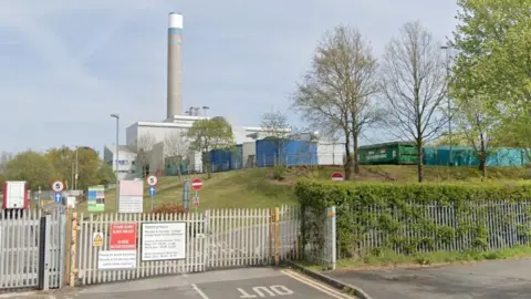 Closed metal gates and a fence surround the incinerator site, with grass beyond and the grey incinerator itself and the tall chimney above it