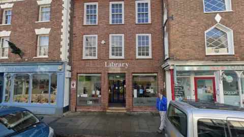 Red brick three story building, with two large, square windows on the ground floor, and a door between them. The word Library is written in large white text above the door. The other two floors have three white-framed windows each. The building is surrounded by two more three-story red brick buildings, which sit slightly forward of the library.