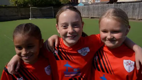 Erin, Elise and Penny are smiling, wearing orange football tops, on a football pitch with a goal in the background. 