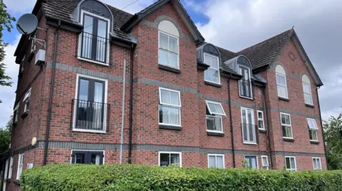 A three-storey block of flats of red-brick with a green hedge in the foreground.