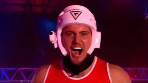 BBC /  Hungry Bear Media Ltd A contestant in Gladiators wearing a white helmet and a red shirt with the logo.