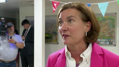 Eluned Morgan wearing a white blouse and pink blazer, looking off to side, with bunting above her head and a man holding a camera in the background