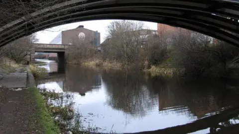 Tinsley Canal in the Darnall area of Sheffield on an overcast day. There's a towpath next to the canal and a factory on the other side of the water.