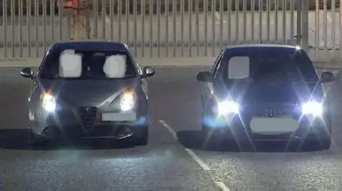 Two cars side by side with their lights on and reg plates blacked out, in what looks like an empty car park at night.