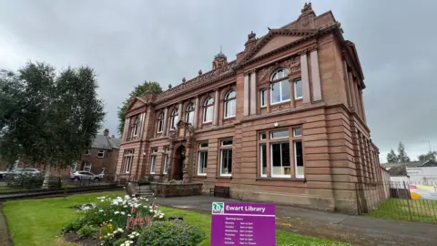 The entire front of the Ewart Public Library from the corner. It's a large red sandstone building with ornate victorian detailing.