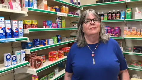 The image shows a woman wearing a blue T-shirt with a tassel necklace. She is in a pharmacy and behind her are rows of medication.