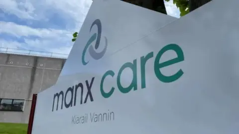 A large white sign with the words Manx Care in English in Manx in grey and green writing on it along with the healthcare provider's logo, which resembles a Celtic knot.