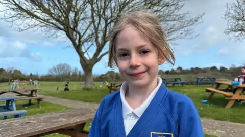 A young girl with blonde hair and brown eyes in a blue school uniform smiles at the camera, with a play field in the background.