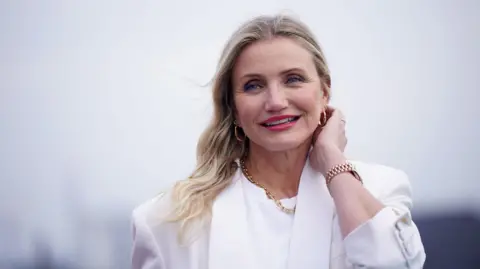 PA Media Cameron Diaz wearing a white blazer and white blouse. Her hair is open and over her right shoulder, and she wears gold hoop earrings and a gold chain necklace.  With her left hand, she is pushing hair behind her ear.