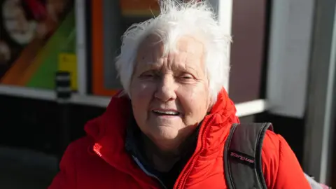 A woman with white hair wearing a red coat and blue jumper, with a black bag strap on her shoulder. She is standing in front of a shop, which can be seen out of focus in the background.