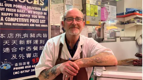A man with short white hair and glasses. He is wearing a white shirt and a dark red apron. He has tattoos on his arms and is leaning his elbow on a fridge.