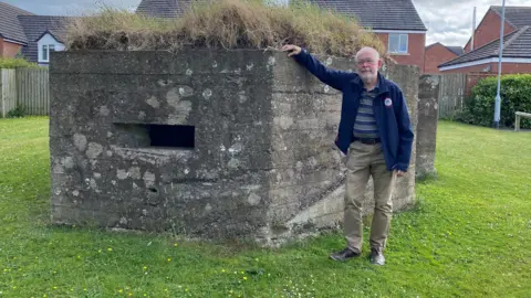 Alan Sture is a man in his 60s wearing brown trousers with a blue jacket. He's standing by a pillbox, a concrete hexagonal defensive structure from the 2nd world war where men could hide and shoot at the enemy from. Behind it are some moden houses.  