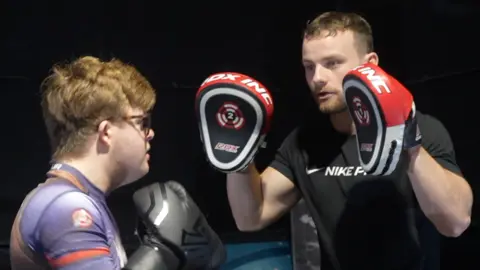 Jamie Niblock/BBC The two men are pictured sparring in a boxing ring. Edward Wagland is wearing boxing gloves while Callum Foster faces his with boxing pads on his hands.