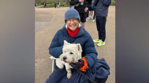 Jacqueline Krarup A woman wearing a navy hat, coat, red scarf and leather gloves sitting in a wheelchair smiling at the camera. She is holding a small white dog on her lap. 