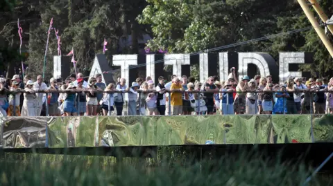 Jim Dyson/Getty Images Crowds of festival-goers lined-up on a bridge overlooking the lake. In the background is the Latitude sign.