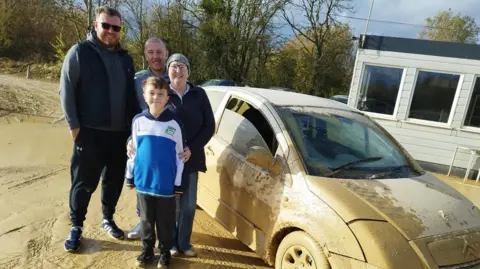Sally Sarl Sally with her two sons and young grandson standing next to a muddy rally car. Sally is wearing a head scarf, jeans and a navy jumper. Her family are all wearing dark tracksuit bottoms and hoodies. 