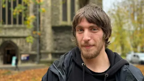 BBC Ryan stands in front of St Mary's Church in Southampton, which is blurred in the background. He has medium length brown hair and a beard. He wears a black hoodie and a black raincoat over the top.