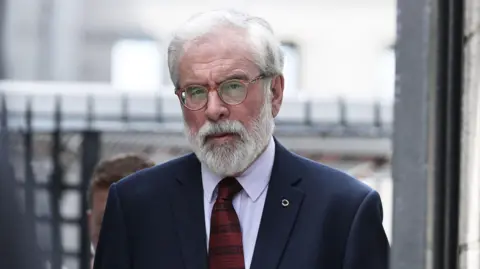 Gerry Adams, pictured entering a courthouse, he has grey hair and a grey beard. Wearing glasses, a white shirt and navy suit jacket, with a striped patterned red tie.
