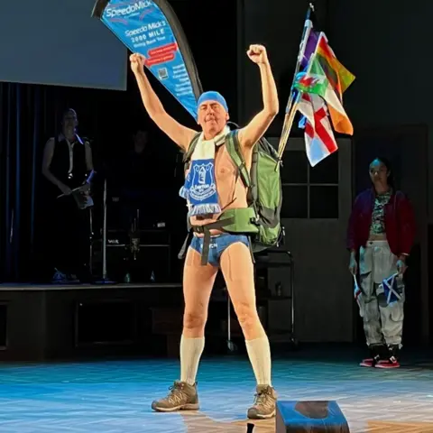 Clara Mbirimi/Liverpool's Royal Court Paul Duckworth as Speedo Mick on stage in a pair of blue SPeedos and Everton scarf with his arms in the air