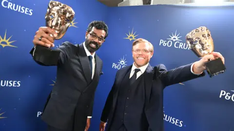 Getty Images Romesh Ranganathan and Rob Beckett in suits holding BAFTA awards.