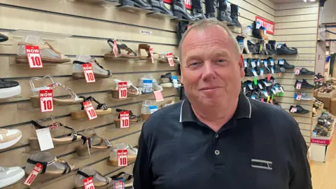 Photograph of Ray Smith at Wigan Indoor Market. The image show him in front of his shoe store. He wears a dark blue polo shirt.