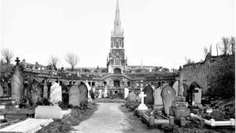 Heritage England A black and white photograph showing a church spire in the centre of the image surrounded by grave stones of different shapes and sizes. Below the church are two levels with openings to the catacombs. 