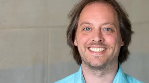 Dr Wijnand van Tilburg is wearing a turquoise shirt in front of a beige wall. he has long brown hair and is smiling.