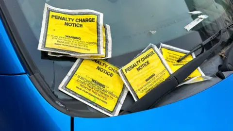 A collection of penalty charge notices in yellow pockets on a blue car's windscreen. 