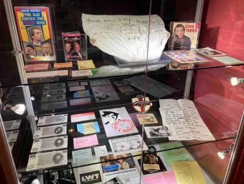 Memorabilia is visible on glass shelves. The top shelf includes recordings of Mayall, the next shelf features tickets and a letter and the bottom area includes photos.