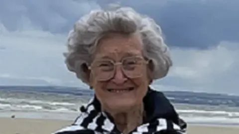 Joyce Taggart is smiling whilst wearing a black and white striped coat . She is standing on the beach.