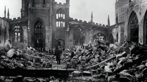 PA Media An old black and white photo showing inside a ruined cathedral with rubble strewn everywhere while the outer portion of the building remains standing. People can be seen entering the site and looking around at the scene.