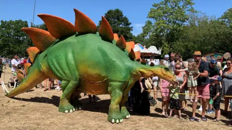 Gloucester BID A large green plastic dinasoar with children surrounding it, petting it.