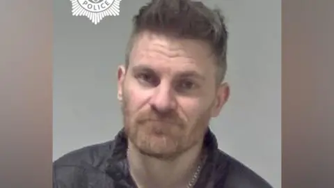 A man with spikey dark grey hair is pictured wearing a black coat in front of a grey/neutral background.