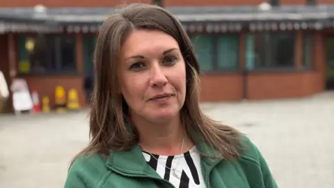 Wearing a green quarter zip jumper and black and white top, Laura Fowler stares at the camera. She has long brown hair which flows down just below her shoulders. The exterior wall of the community centre can be seen in the background.