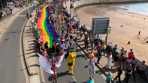 Hundreds of people walking along Torquay's seafront and next to a beach as they take part in the march for Torbay Pride in 2023. A large rainbow flag has been unfurled and is stretching back across the crowd. There are a number of people in fancy dress and a woman wearing rainbow clothing walking on stilts.