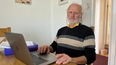 Chris Feast sits at his wooden dining table with a hand resting on an open laptop computer, and paperwork to his right. He is wearing glasses, a black jumper with grey and cream stripes, and a yellow shirt.