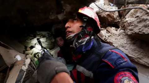 A firefighter wearing a helmet and heavy-duty jacket holds a small camera device up in his hand as he searches for survivors of the earthquakes in La Guaira, Venezuela.