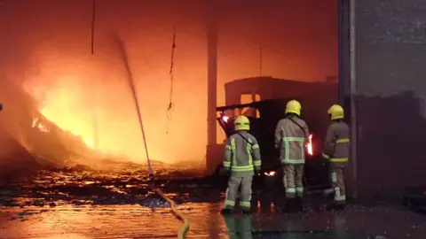 Three firefighters wearing beige protective kit and yellow helmets standing with their backs to the camera and facing a large fire in a waste building.