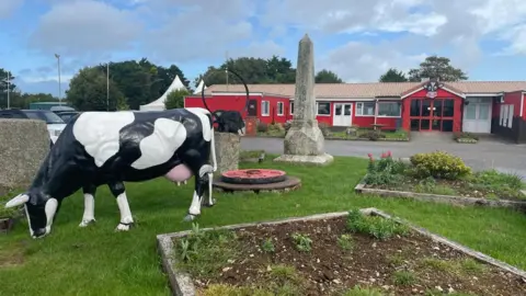 The entrance to Dairyland. It features model black and white cow on grass in front of a red building, with a large wooden cow's head above the building's entrance. There is also a stone obelisk in front of the building.