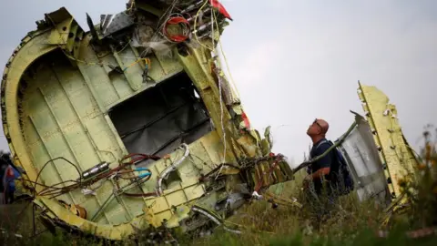 A Malaysian air crash investigator inspects the crash site of Malaysia Airlines Flight MH17 in the Donetsk region of Ukraine in July 2014.  The man is looking at the wreckage of a plane, which has lots of exposed wires. 