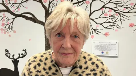 Community worker Jeanette Warke has short white hair and is wearing a leopard print cardigan and white top. She is standing near a mural of a tree and deer.