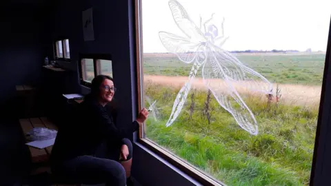 Steart Marshes Dragonfly Trail A woman is sitting on a bench in a building which has a few very small windows and one very large window. On the large window the woman is painting a big dragonfly in what appears to be white paint. Grass fields can be seen outside. The woman is smiling and looking at the camera.