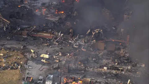 An aerial view of an industrial sit covered in debris. There are flames dotted throughout the site and black smoke rising.